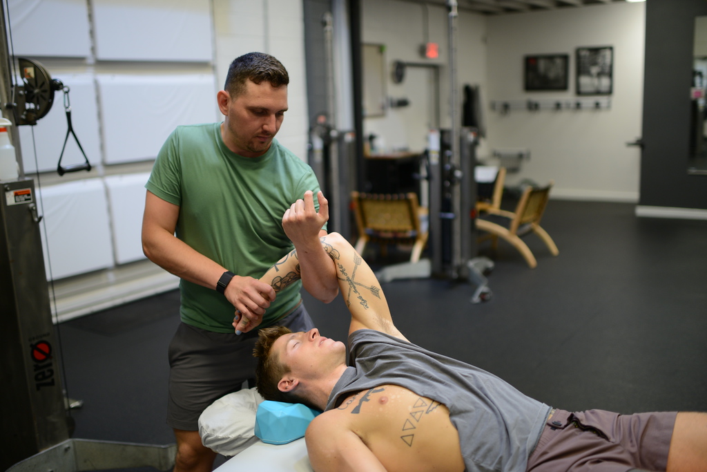 Chris supports a client's raised arm while the client rests on the table in athletic clothing.